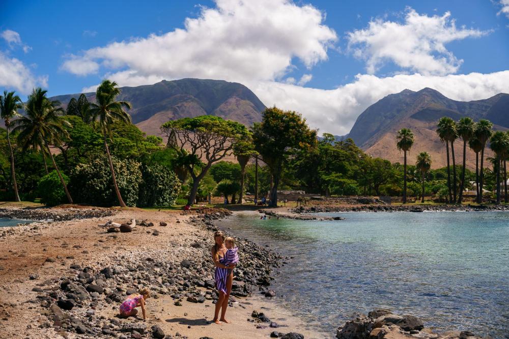Người phụ nữ và trẻ em trên bãi biển đá tại Maui với dãy núi phía xa