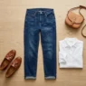 Bộ trang phục casual với quần jeans tối màu, áo trắng, giày lười và túi đeo chéo trên nền gỗ